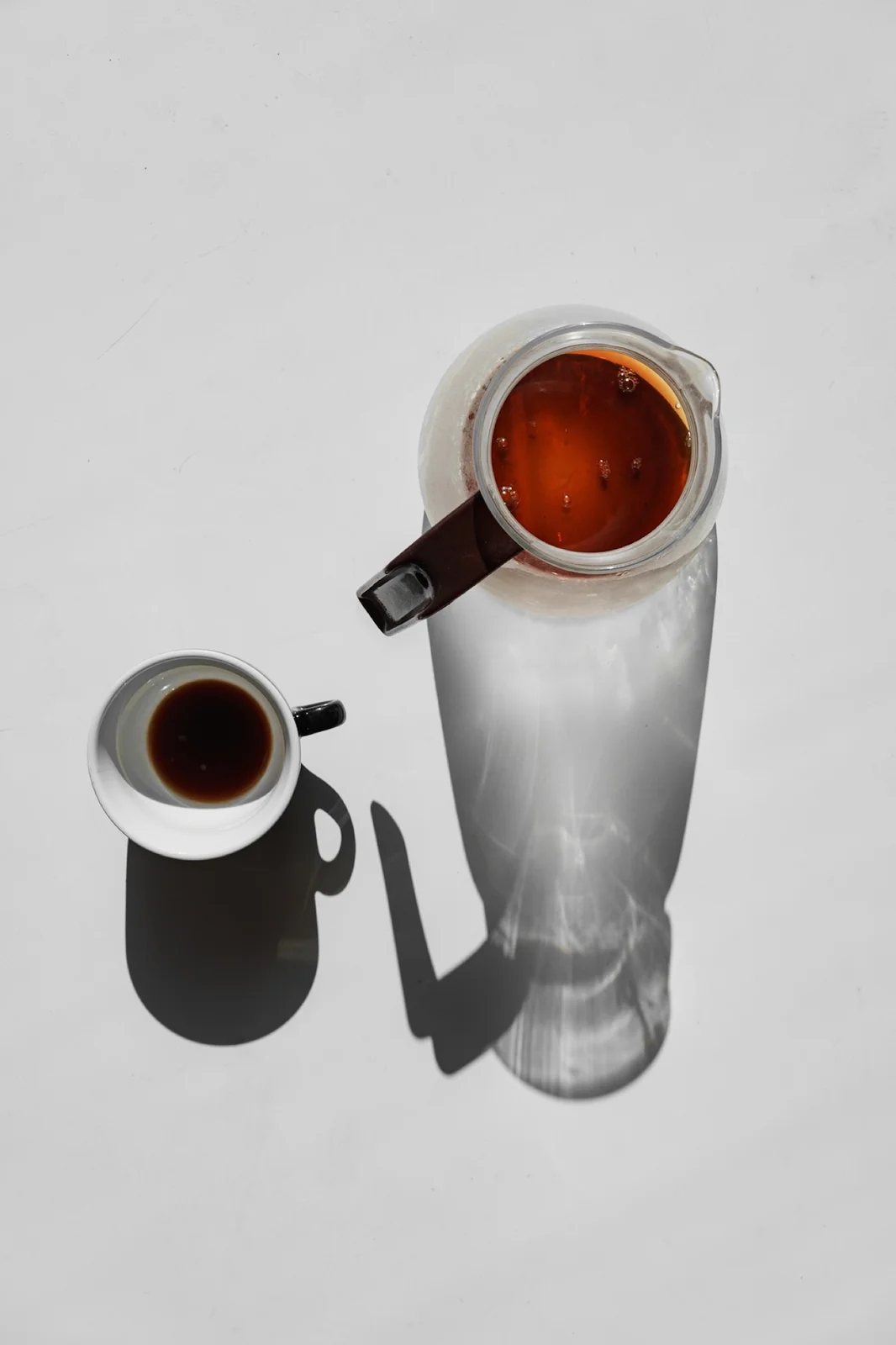 A glass jug and a small coffee cup cast long shadows across a pale surface.