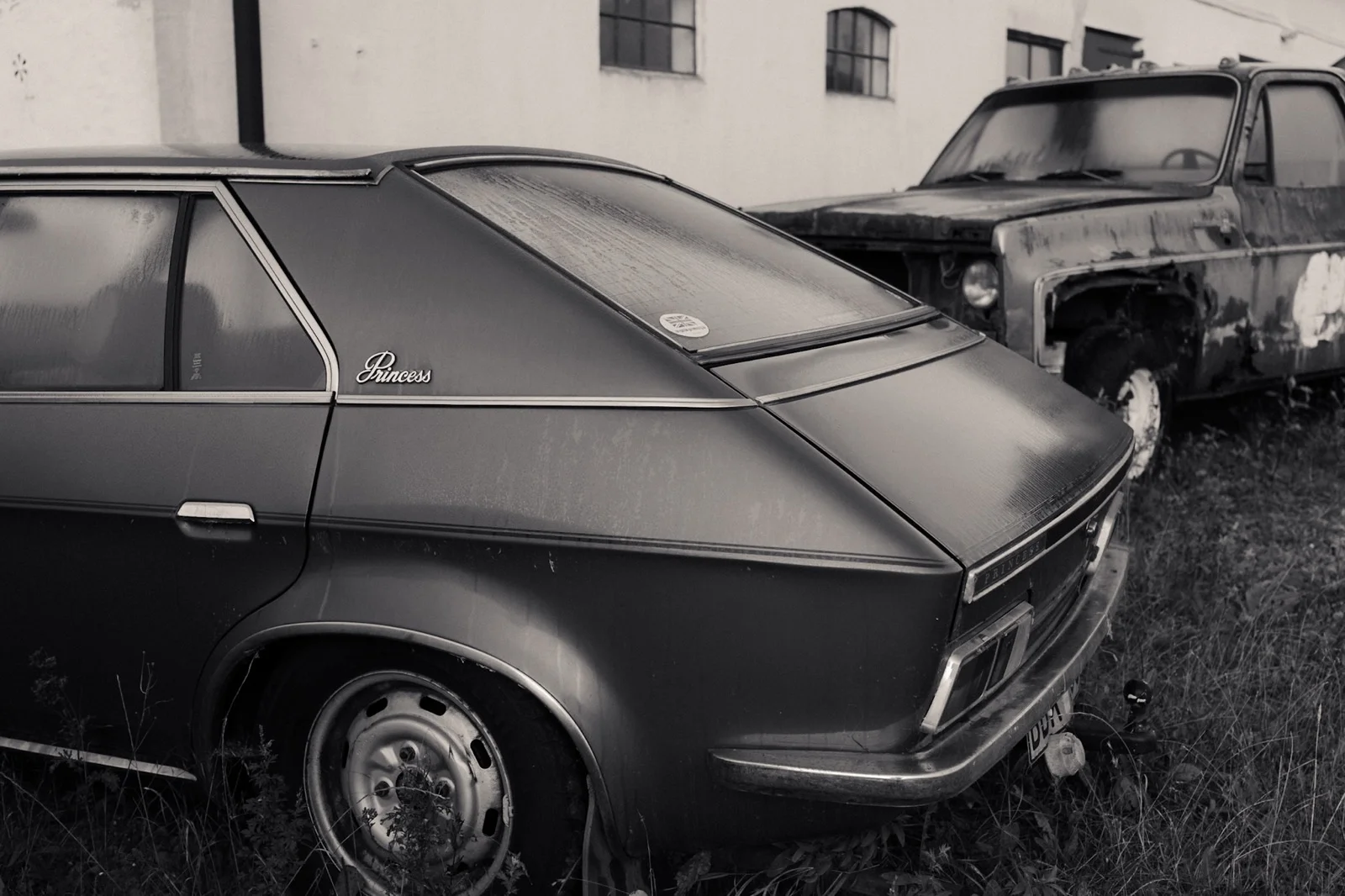 A black-and-white photograph of two weathered cars parked beside a building.