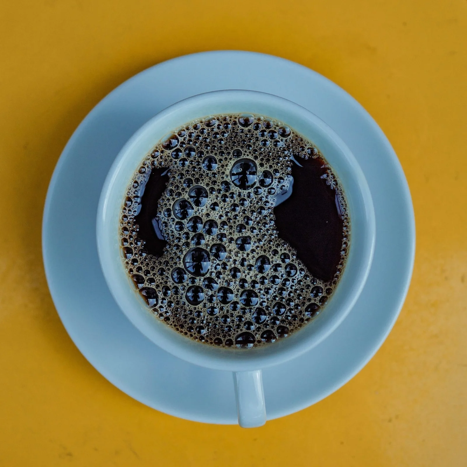 A blue cup of black coffee with bubbles on a bright yellow surface.