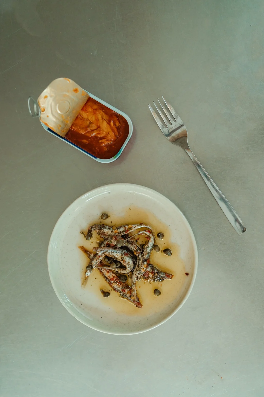 A bowl of food beside an open tin and a fork on a grey-green tabletop.